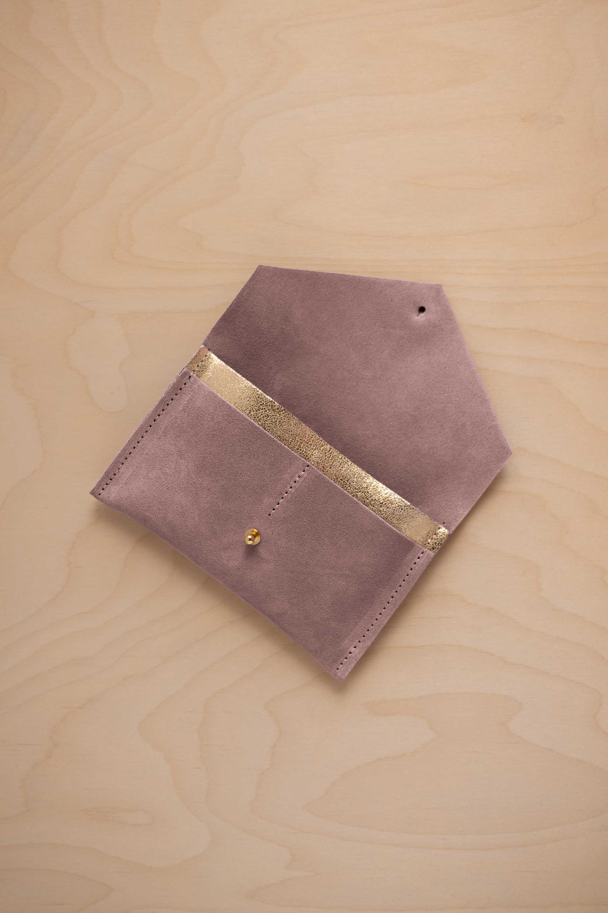 A dusty mauve grey suede pouch with a metallic gold contrast pocket and a small gold stud, displayed on a light wood grain background.