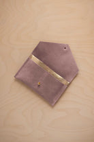 A dusty mauve grey suede pouch with a metallic gold contrast pocket and a small gold stud, displayed on a light wood grain background.