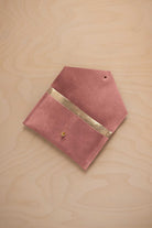 A dusty rose pink coloured suede pouch with a metallic gold contrast pocket and a small gold stud, displayed on a light wood grain background.