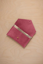 A faded rose pink suede pouch with a metallic gold contrast pocket and a small gold stud, displayed on a light wood grain background.