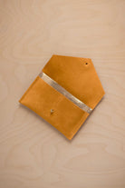 A golden ochre mustard yellow suede pouch with a metallic gold contrast pocket and a small gold stud, displayed on a light wood grain background.