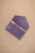 A lavender suede pouch with a metallic gold contrast pocket and a small gold stud, displayed on a light wood grain background.