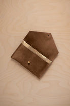 A mocha brown suede pouch with a metallic gold contrast pocket and a small gold stud, displayed on a light wood grain background.
