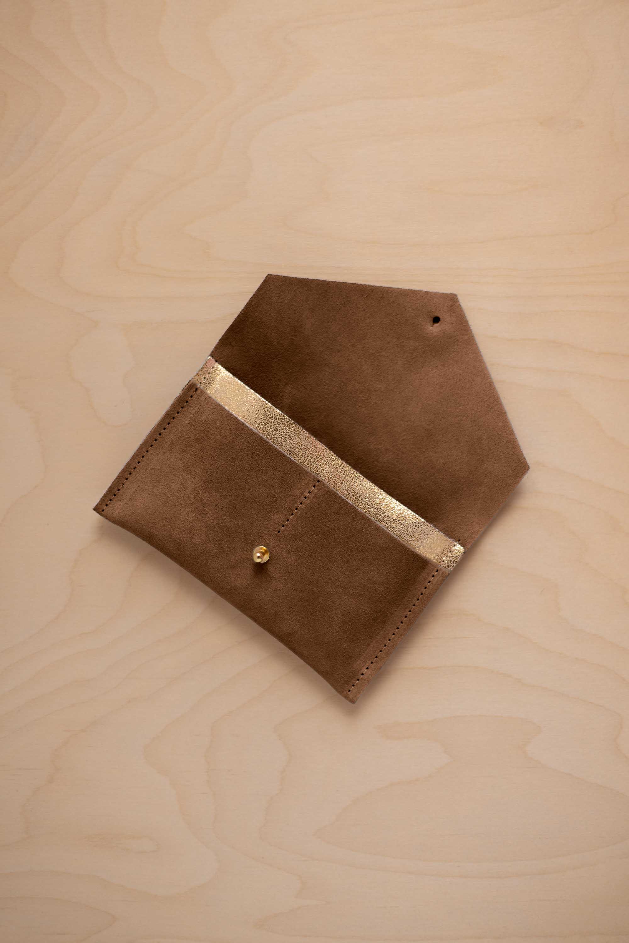 A mocha brown suede pouch with a metallic gold contrast pocket and a small gold stud, displayed on a light wood grain background.