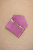 A peony pink suede pouch with a metallic gold contrast pocket and a small gold stud, displayed on a light wood grain background.