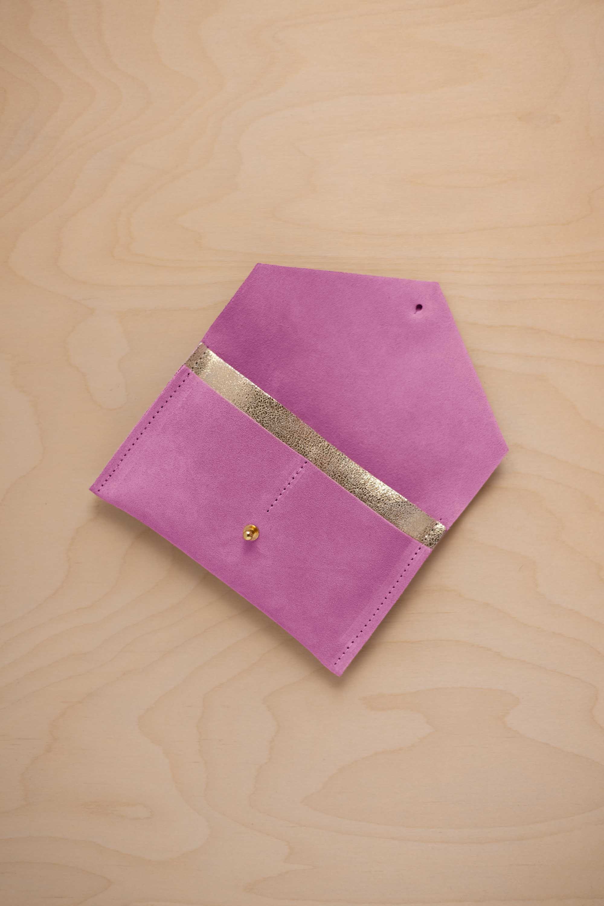 A peony pink suede pouch with a metallic gold contrast pocket and a small gold stud, displayed on a light wood grain background.