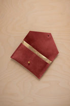 A rust red suede pouch with a metallic gold contrast pocket and a small gold stud, displayed on a light wood grain background.