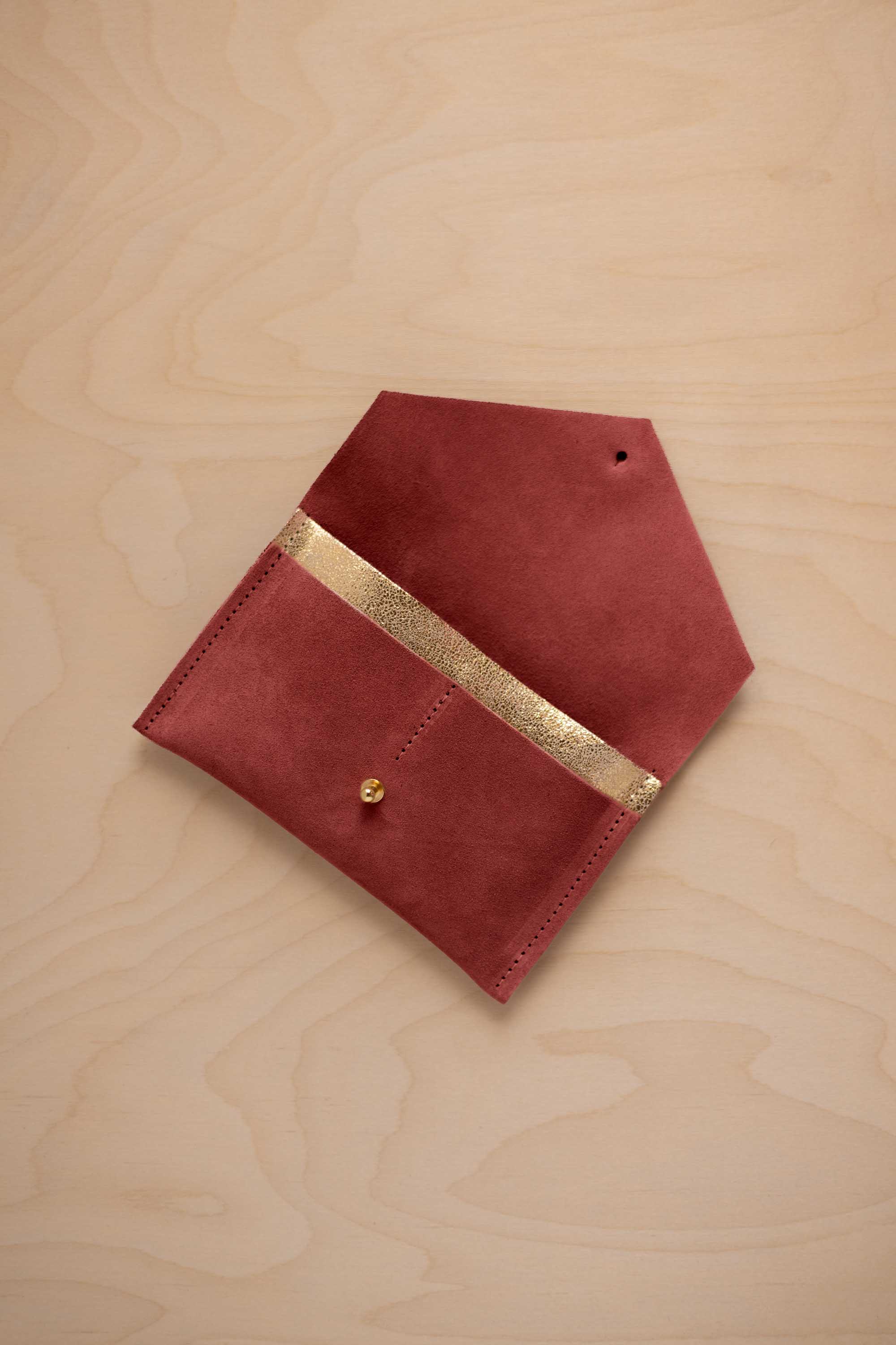 A rust red suede pouch with a metallic gold contrast pocket and a small gold stud, displayed on a light wood grain background.
