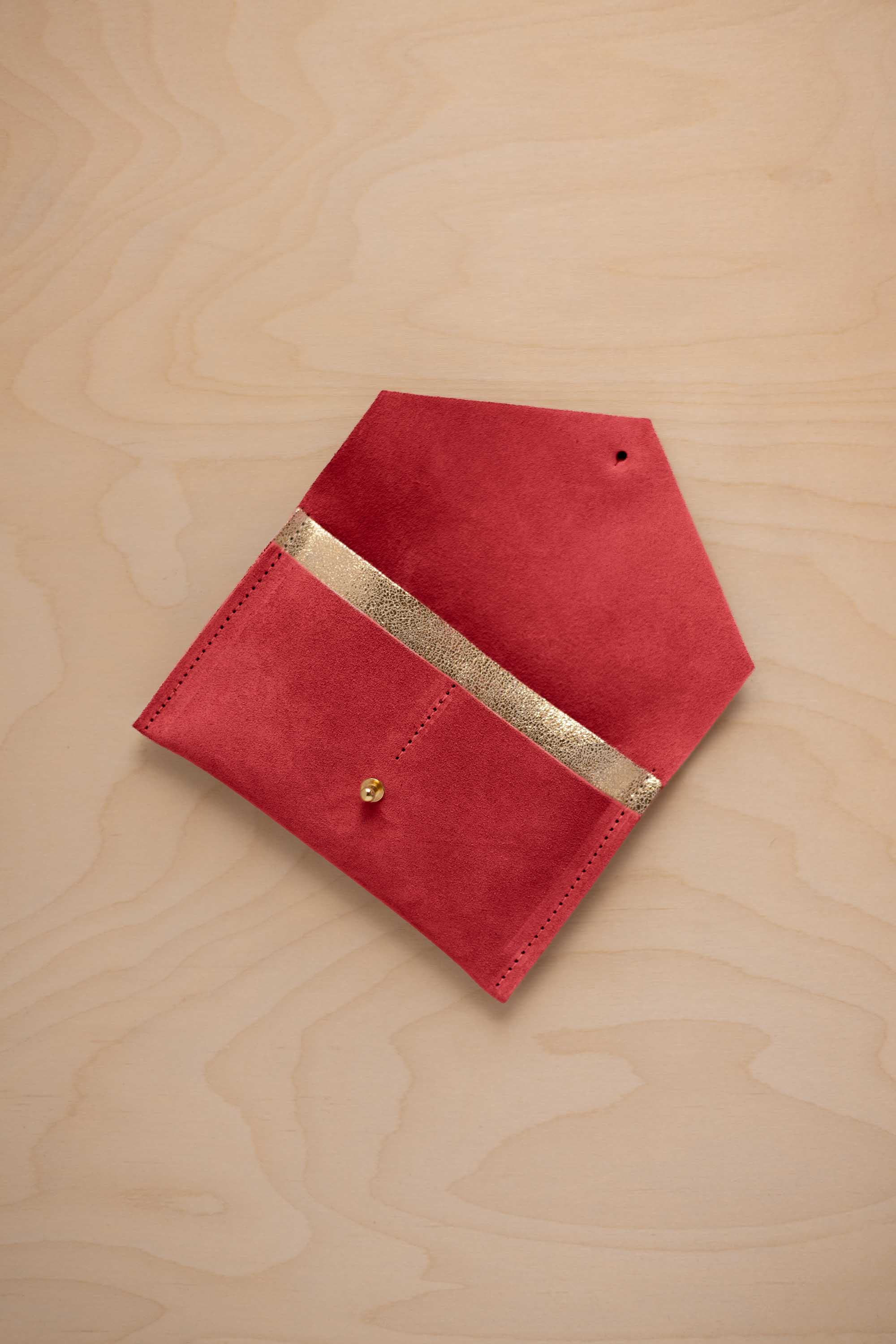 A scarlet red suede pouch with a metallic gold contrast pocket and a small gold stud, displayed on a light wood grain background.