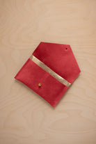 A scarlet red suede pouch with a metallic gold contrast pocket and a small gold stud, displayed on a light wood grain background.