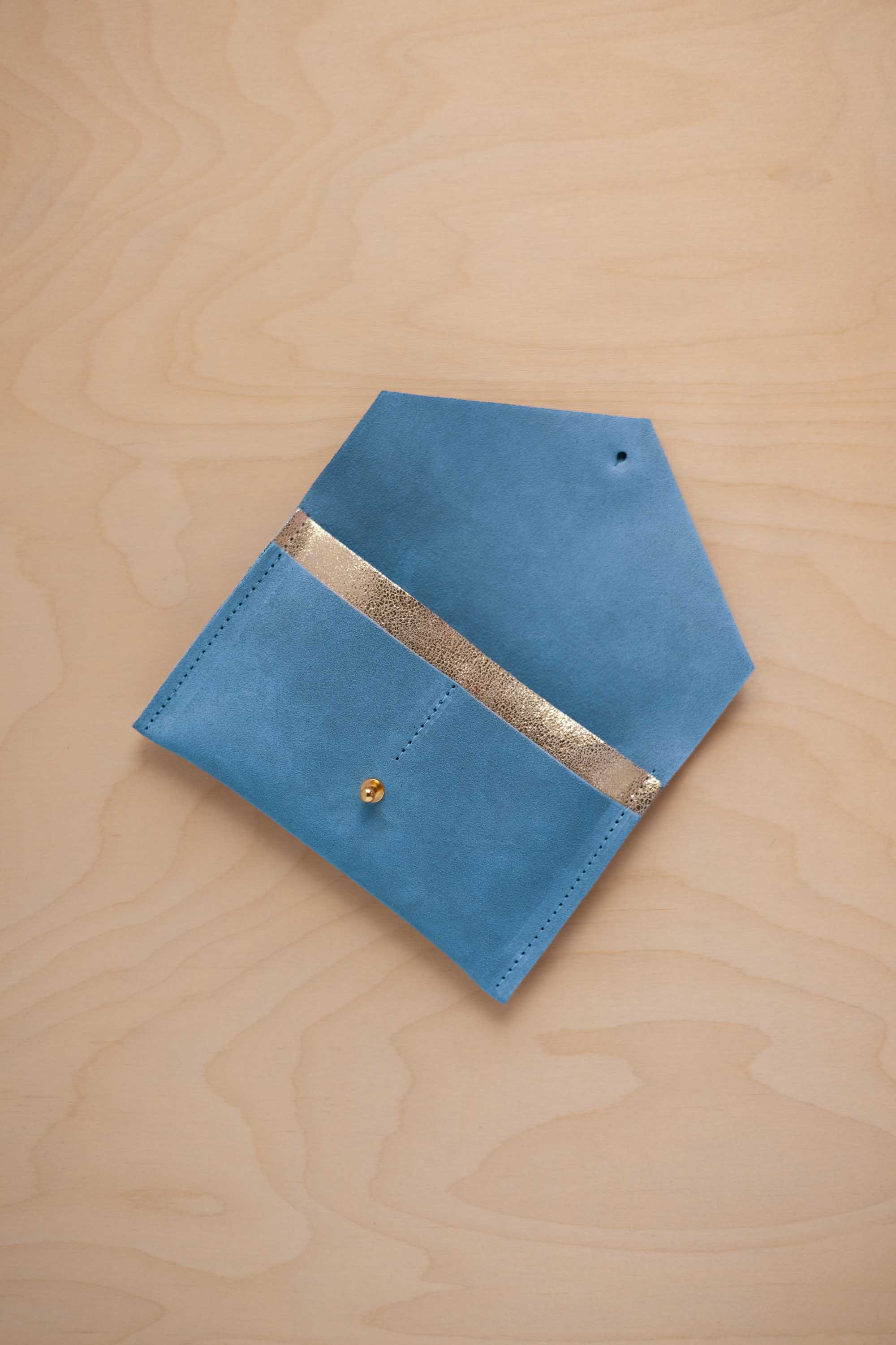 A sky blue suede pouch with a metallic gold contrast pocket and a small gold stud, displayed on a light wood grain background.