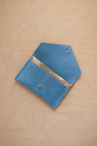 A sky blue suede pouch with a metallic gold contrast pocket and a small gold stud, displayed on a light wood grain background.
