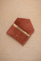 A terracotta suede pouch with a metallic gold contrast pocket and a small gold stud, displayed on a light wood grain background.