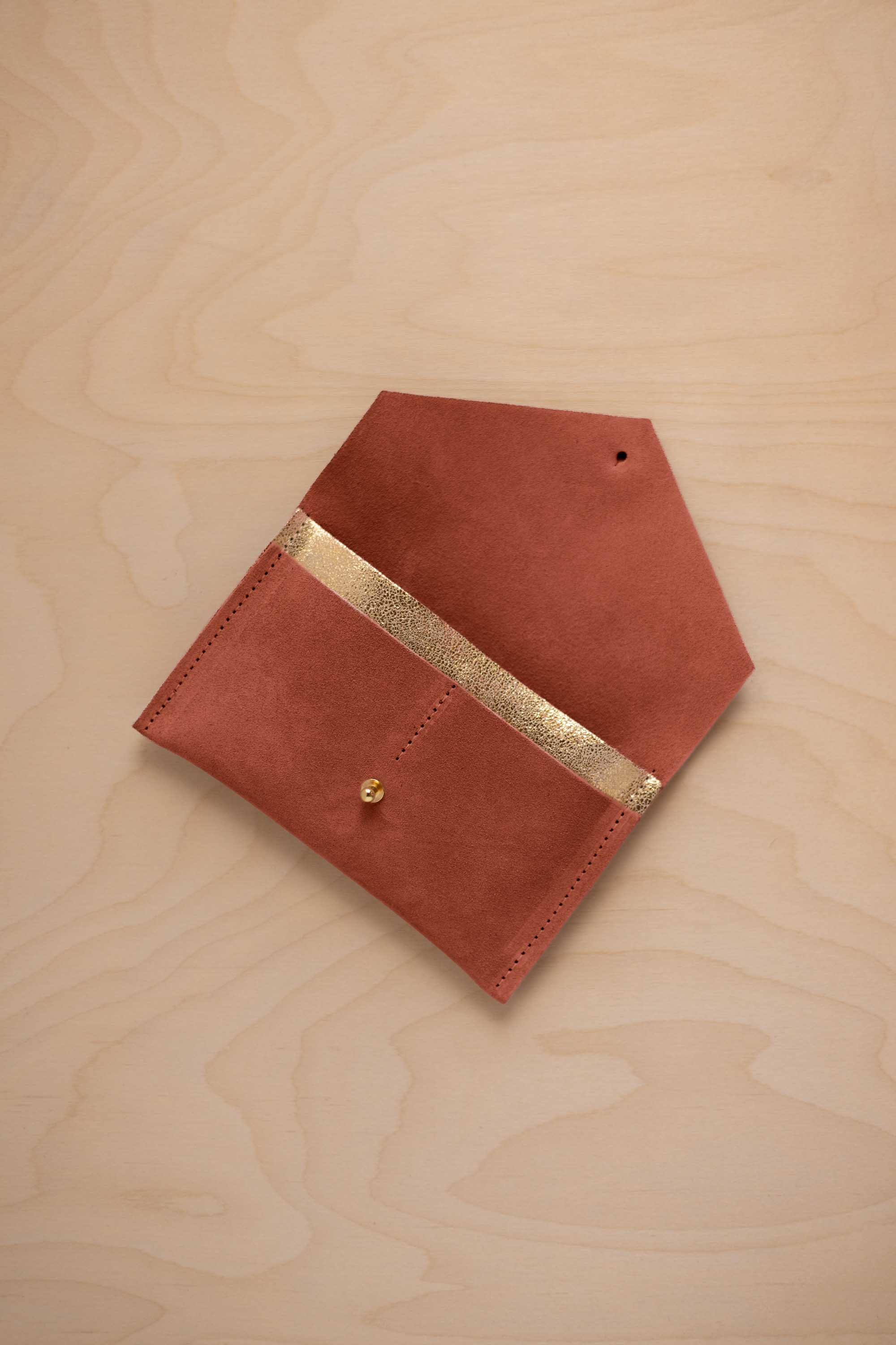 A terracotta suede pouch with a metallic gold contrast pocket and a small gold stud, displayed on a light wood grain background.