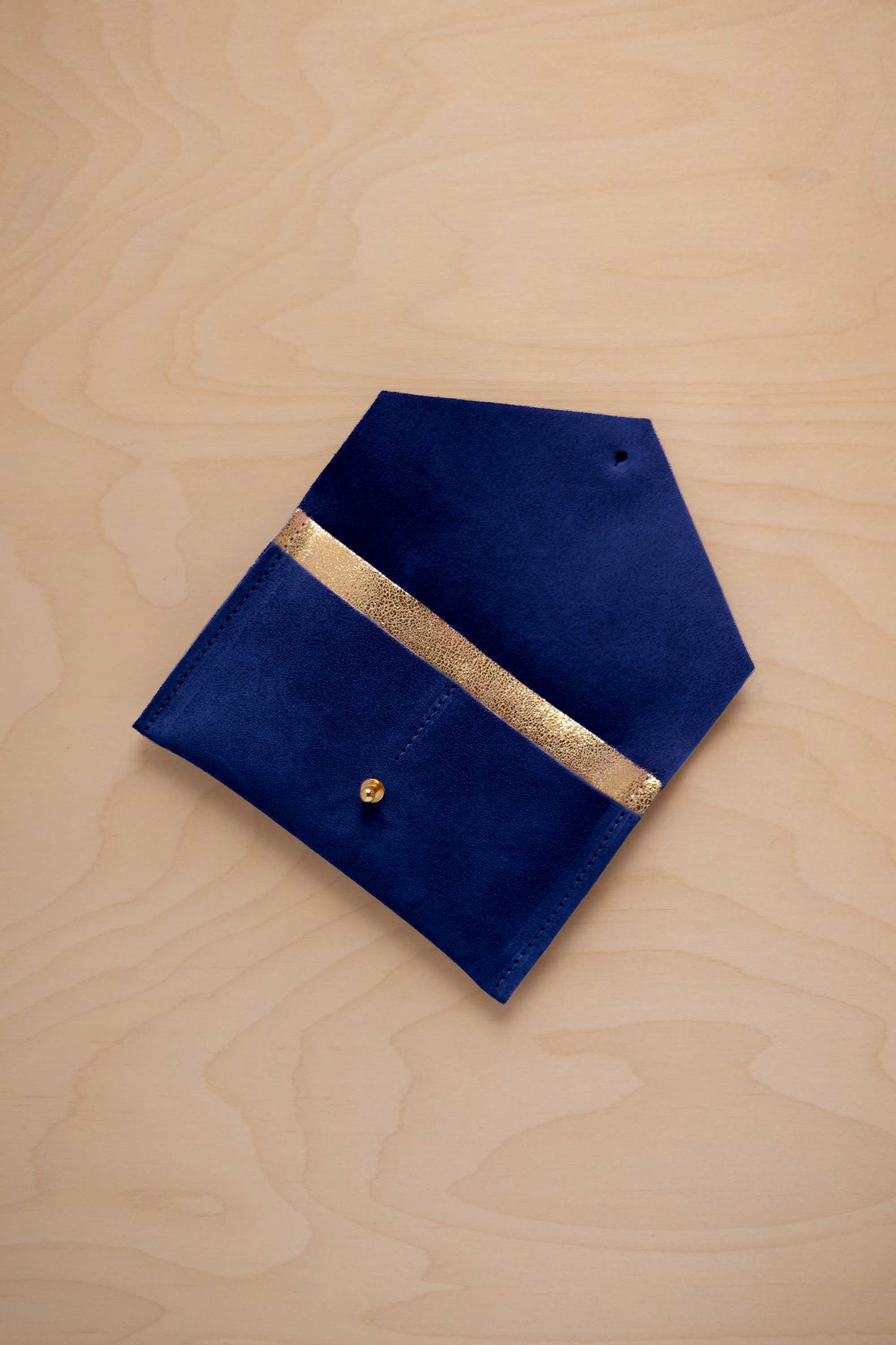 An ultramarine blue suede pouch with a metallic gold contrast pocket and a small gold stud, displayed on a light wood grain background.