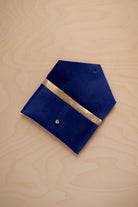 An ultramarine blue suede pouch with a metallic gold contrast pocket and a small gold stud, displayed on a light wood grain background.