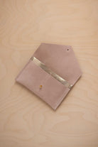 A warm beige suede pouch with a metallic gold contrast pocket and a small gold stud, displayed on a light wood grain background.