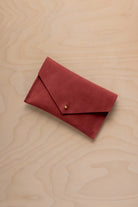 A small red suede envelope-style pouch with a gold snap button, placed on a light wood grain surface.