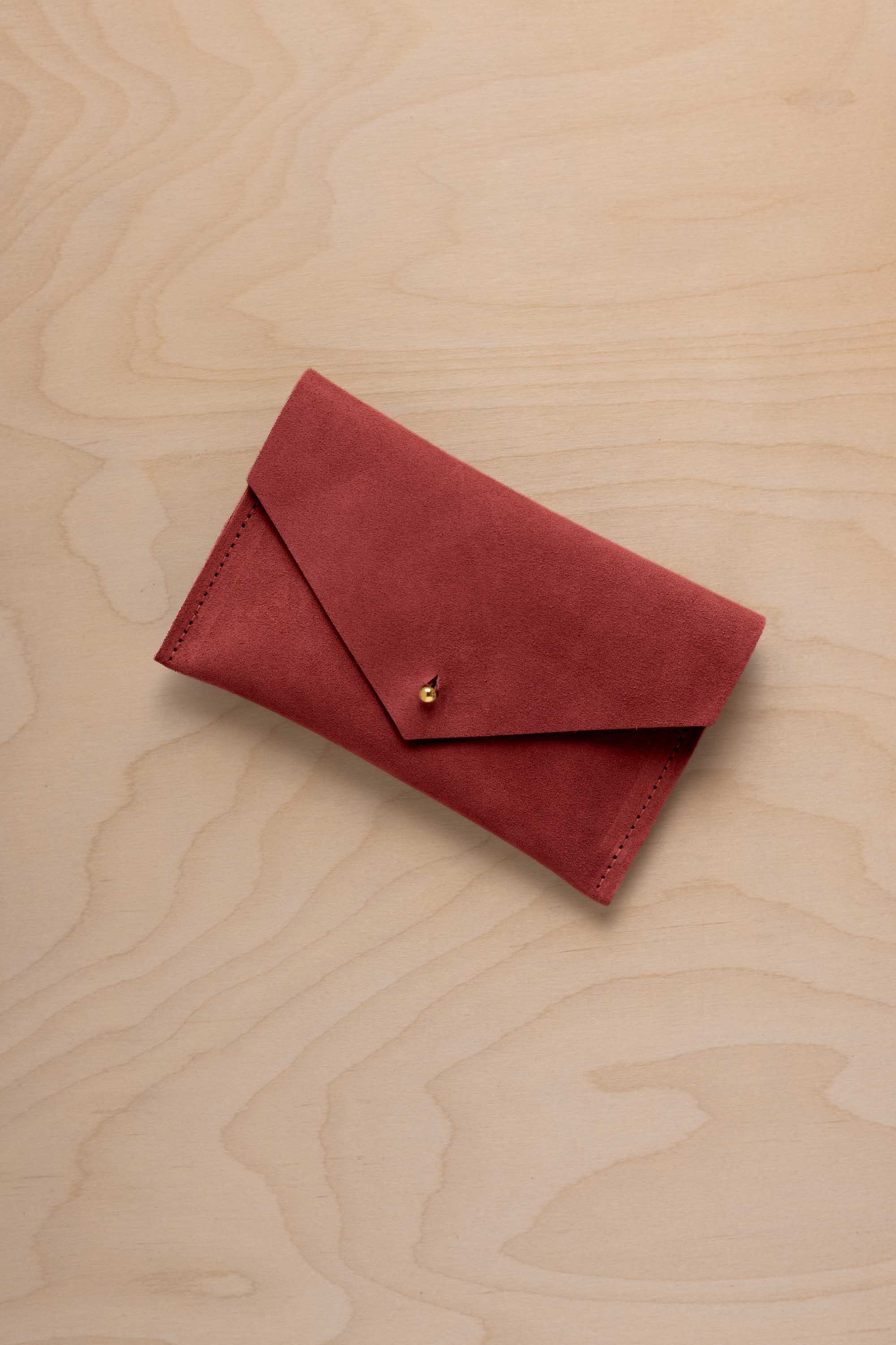A small red suede envelope-style pouch with a gold snap button, placed on a light wood grain surface.