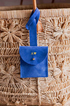 A personalised blue suede luggage tag pouch with the intial "A" on it, a strap and gold button, hanging from a leather handle on a woven straw bag.
