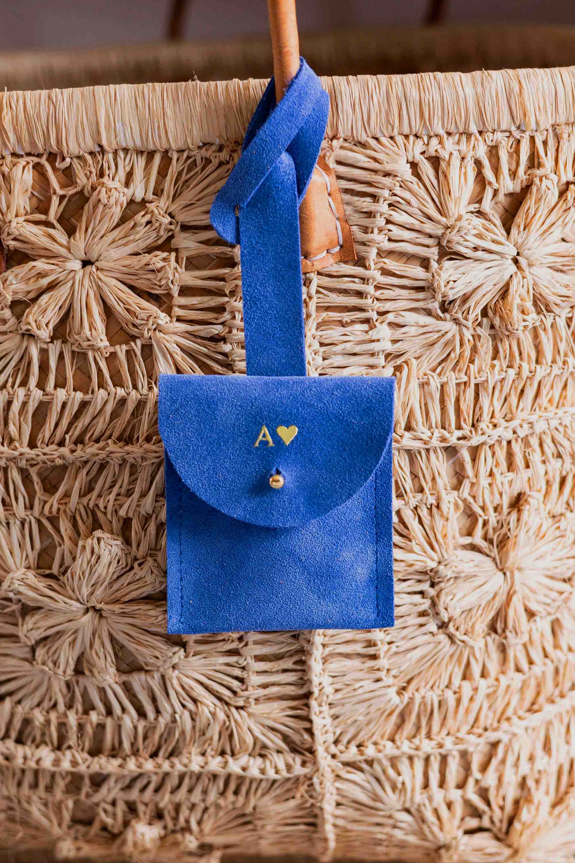A personalised blue suede luggage tag pouch with the intial "A" on it, a strap and gold button, hanging from a leather handle on a woven straw bag.
