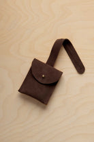 A small espresso brown suede pouch with a flap closure and a matching strap, resting on a light wood grain surface.