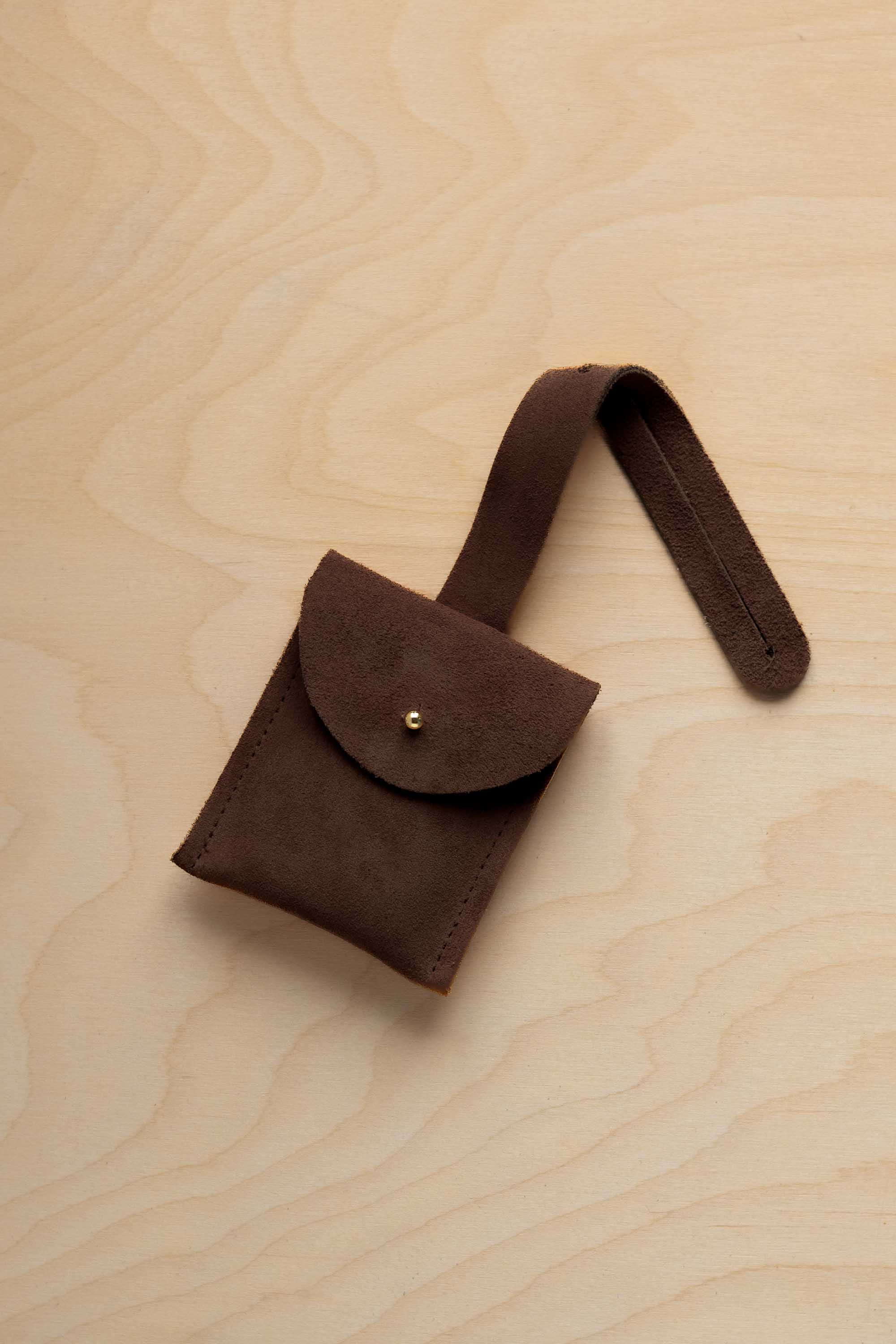 A small espresso brown suede pouch with a flap closure and a matching strap, resting on a light wood grain surface.
