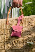 A close-up of a large woven straw tote bag with brown leather handles, featuring a small pink suede luggage tag pouch hanging from one handle. A person's hand with a ring is holding the bag, and a green and white striped dress is visible in the background.