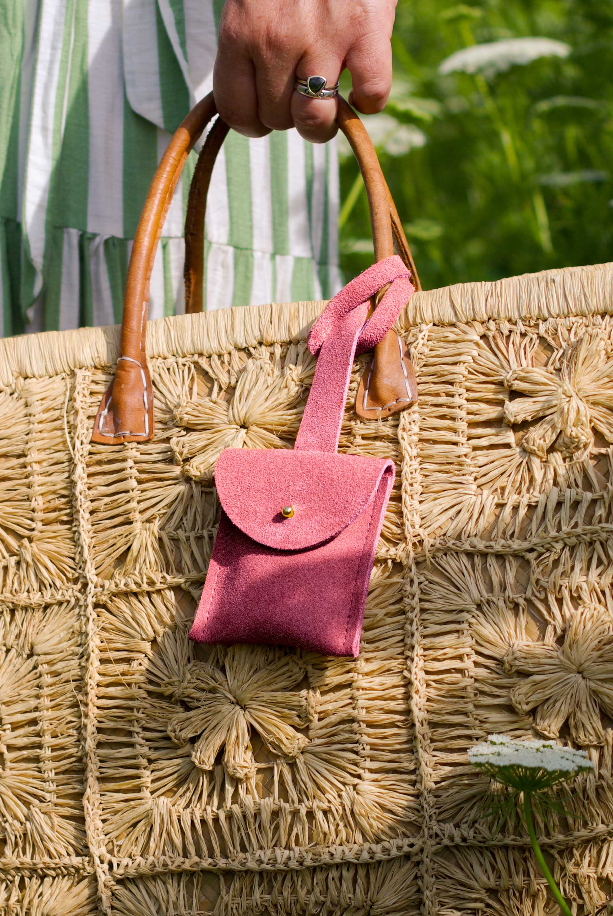 A close-up of a large woven straw tote bag with brown leather handles, featuring a small pink suede luggage tag pouch hanging from one handle. A person's hand with a ring is holding the bag, and a green and white striped dress is visible in the background.