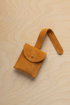 A small golden ochre mustard yellow coloured suede pouch with a flap closure and a matching strap, resting on a light wood grain surface.