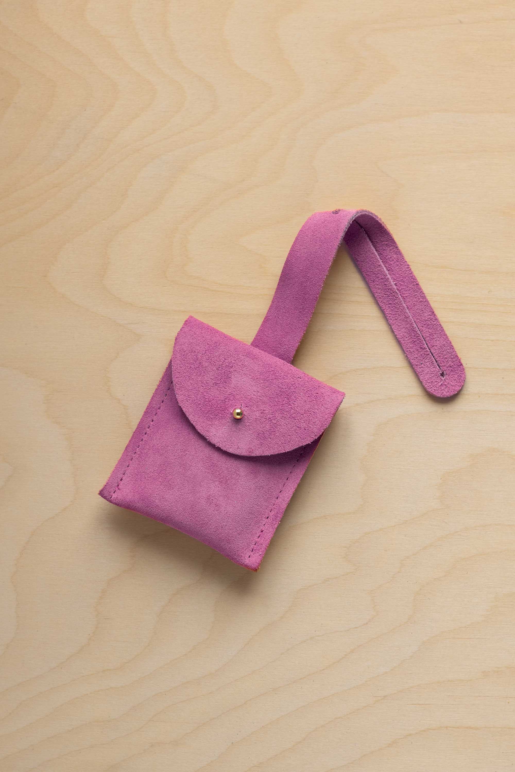 A small pink suede pouch with a flap closure and a matching strap, resting on a light wood grain surface.