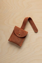 A small terracotta coloured suede pouch with a flap closure and a matching strap, resting on a light wood grain surface.