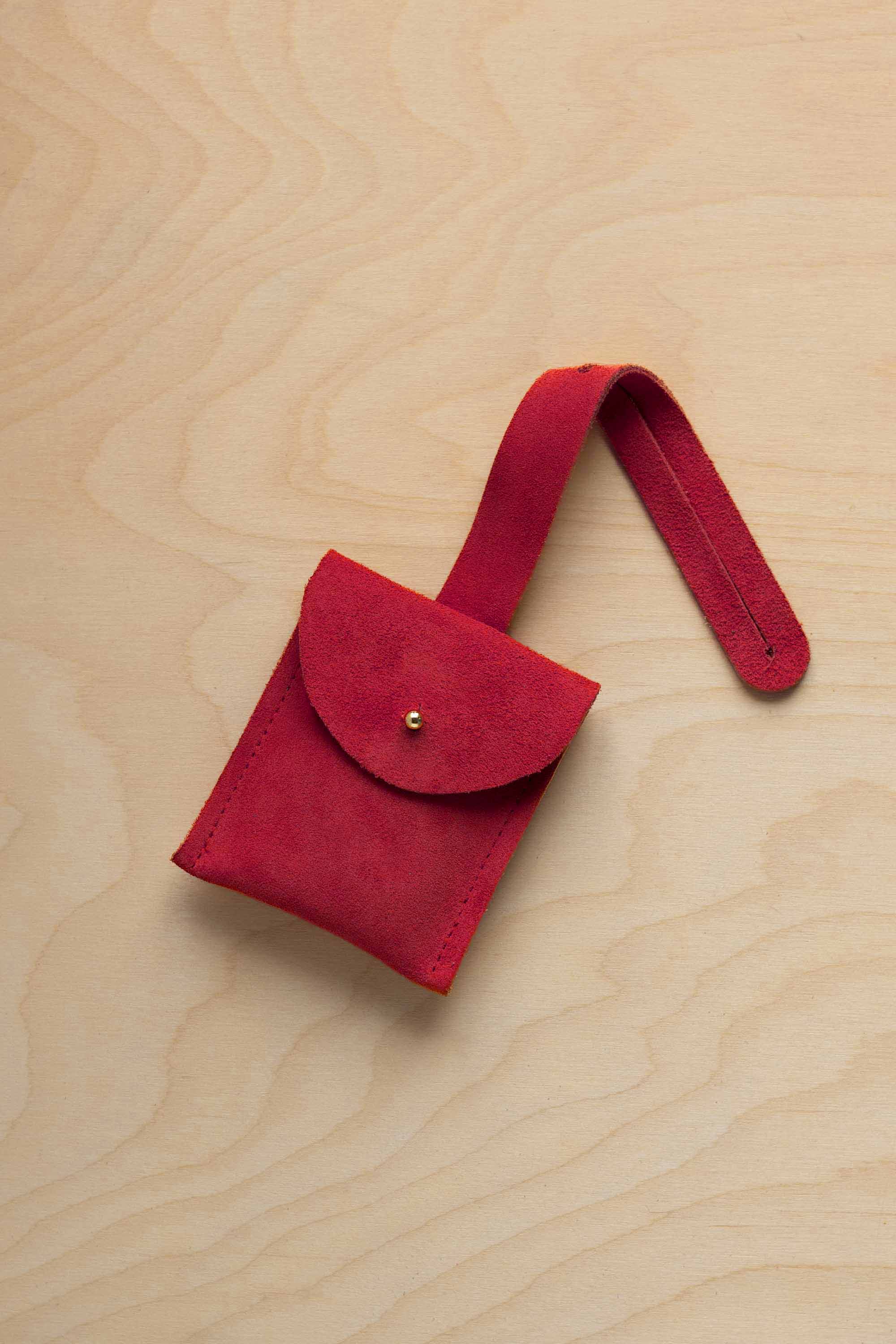 A small crimson red coloured suede pouch with a flap closure and a matching strap, resting on a light wood grain surface.