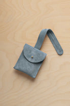 A small dusly blue suede pouch with a flap closure and a matching strap, resting on a light wood grain surface.