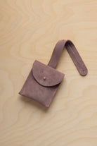 A small dusty mauve grey suede pouch with a flap closure and a matching strap, resting on a light wood grain surface.