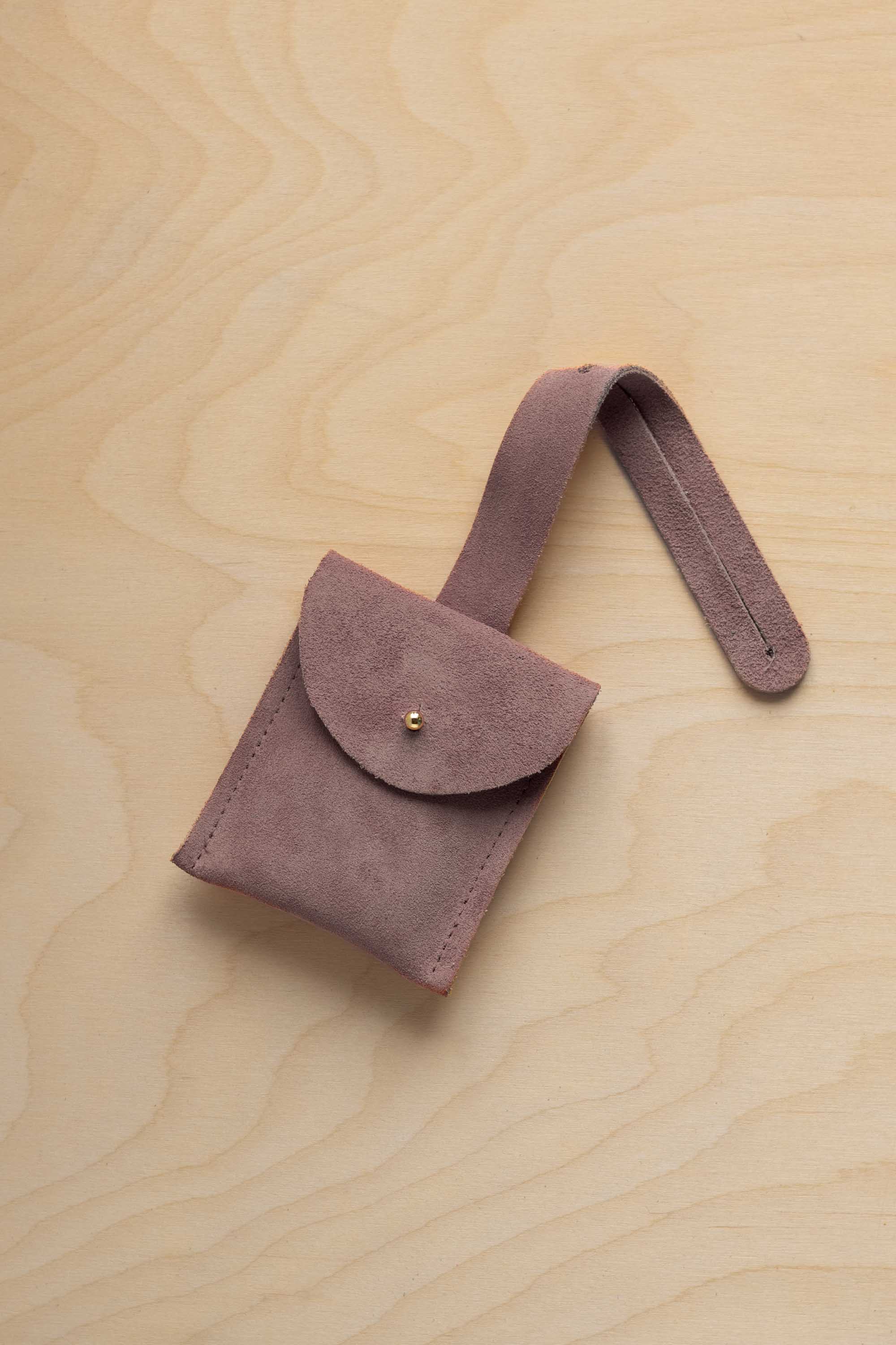 A small dusty mauve grey suede pouch with a flap closure and a matching strap, resting on a light wood grain surface.