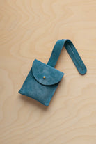A small marine teal suede pouch with a flap closure and a matching strap, resting on a light wood grain surface.