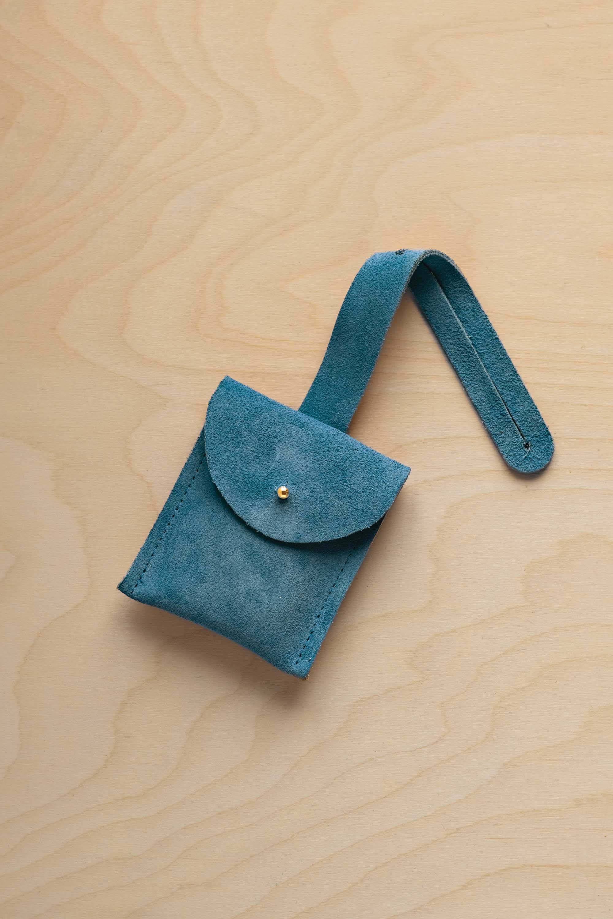 A small marine teal suede pouch with a flap closure and a matching strap, resting on a light wood grain surface.