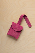 A small raspberry pink suede pouch with a flap closure and a matching strap, resting on a light wood grain surface.