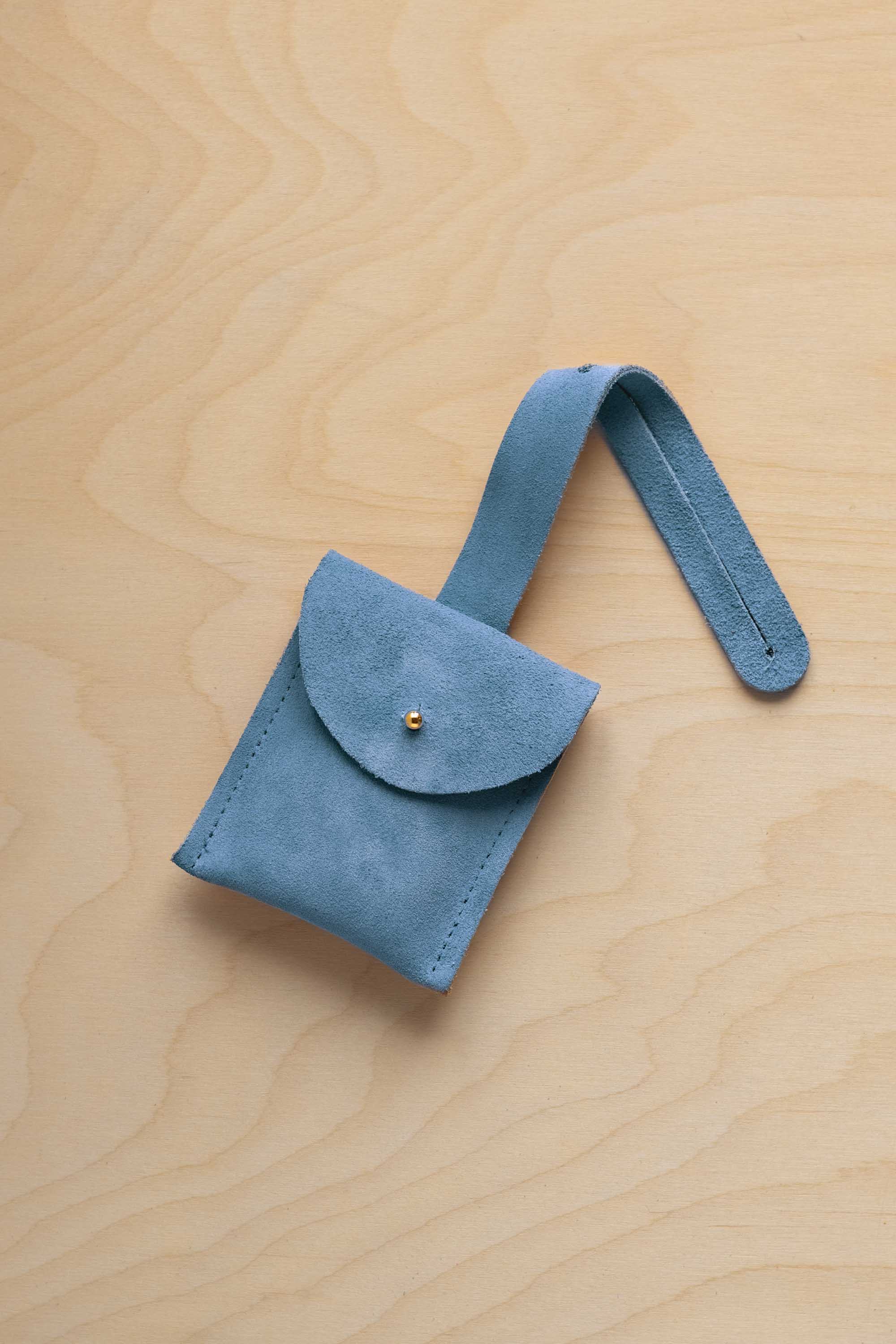 A small sky blue suede pouch with a flap closure and a matching strap, resting on a light wood grain surface.