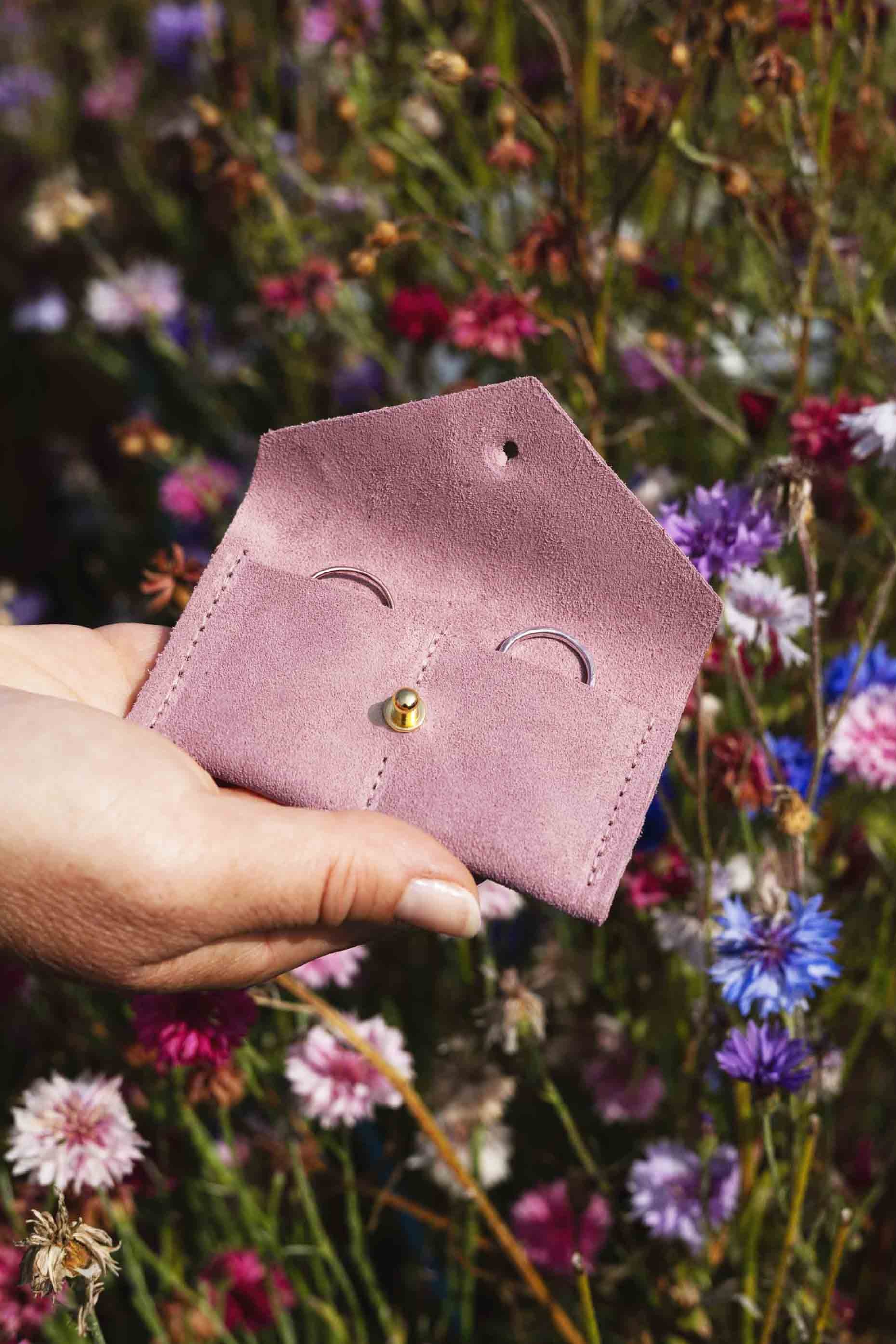 A hand holding a small dusty pink leather pouch with a gold button, set against a blurred background of colourful wildflowers.