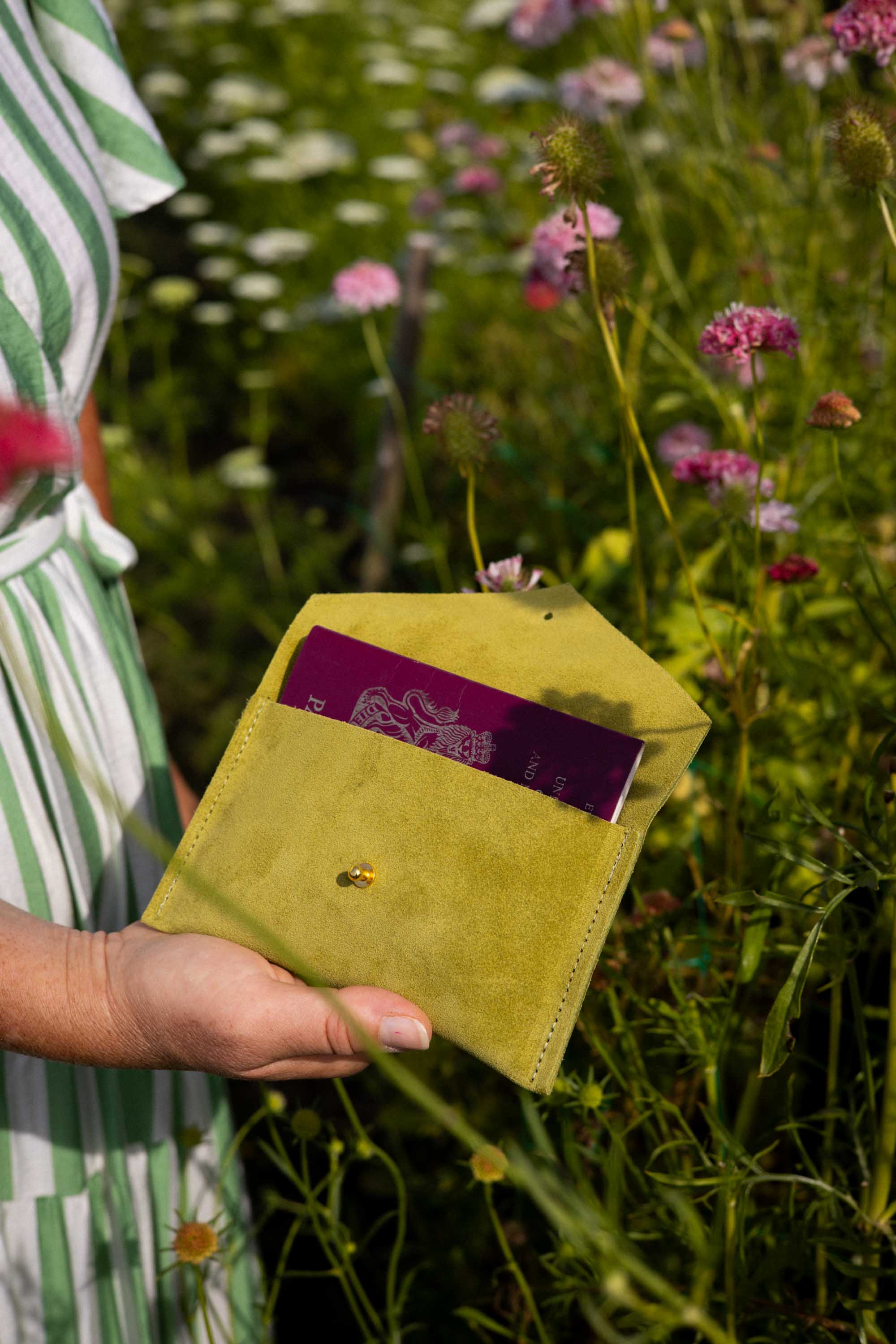 A person outdoors in a flower field holds a small olive-yellow envelope-style pouch with a passport partially inserted. The background is a blurred green and pink wildflower meadow.
