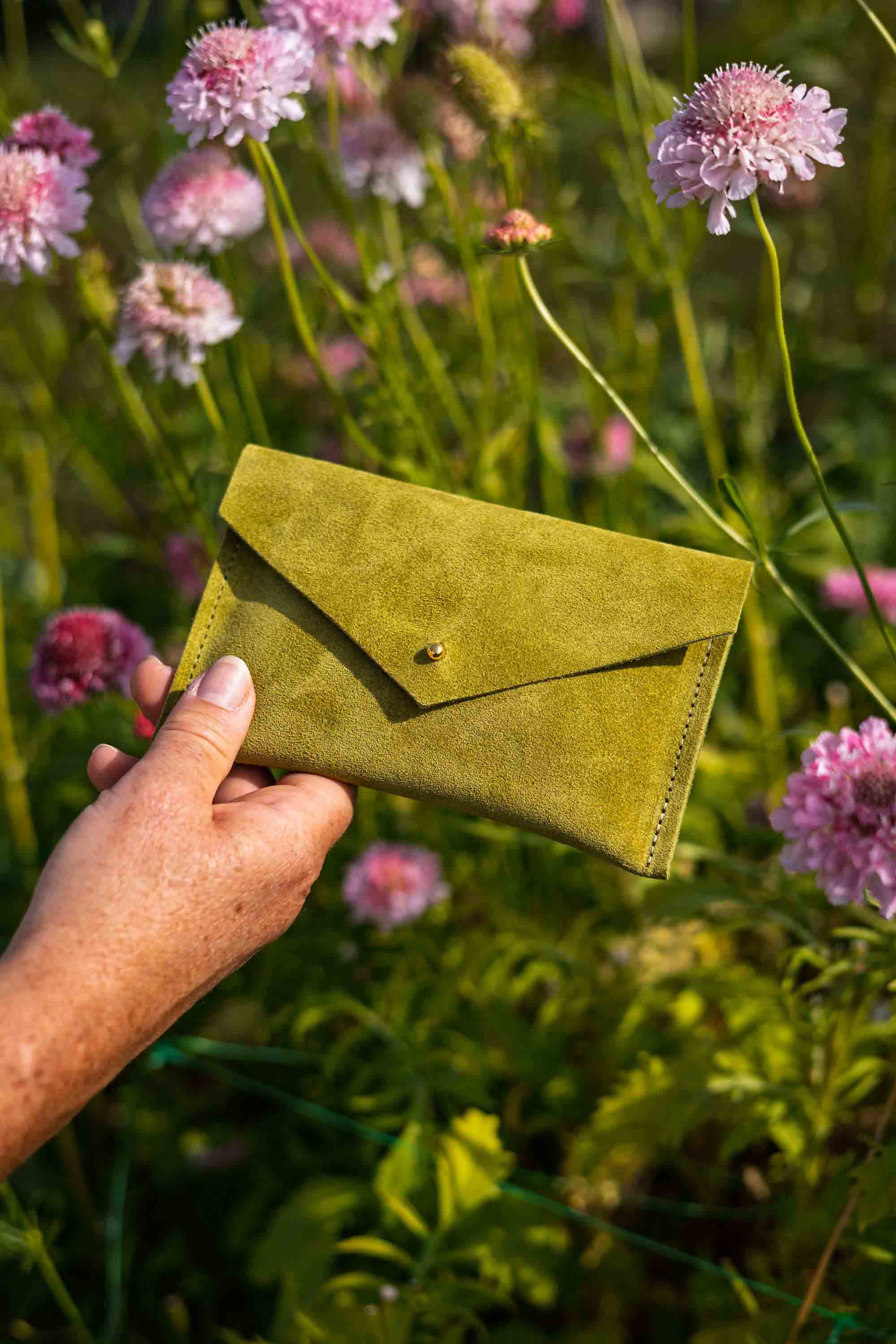 A hand holds a small olive green suede envelope-style wallet or clutch with a button closure, set against a blurred outdoor background of pink flowers and green foliage.