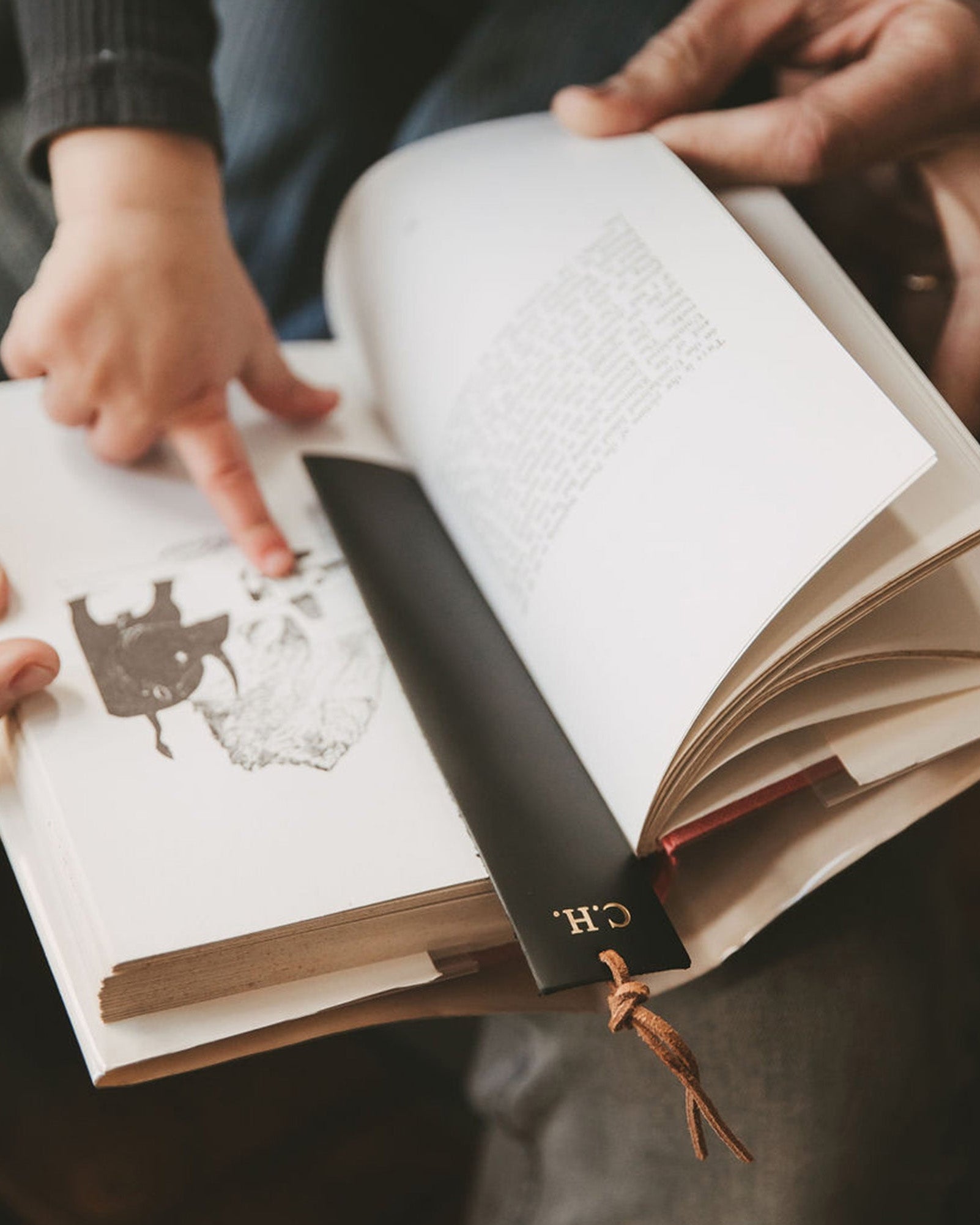 Blackk leather bookmark with monogram "C.H." on it. Held by a baby with his dad whilst reading a book.