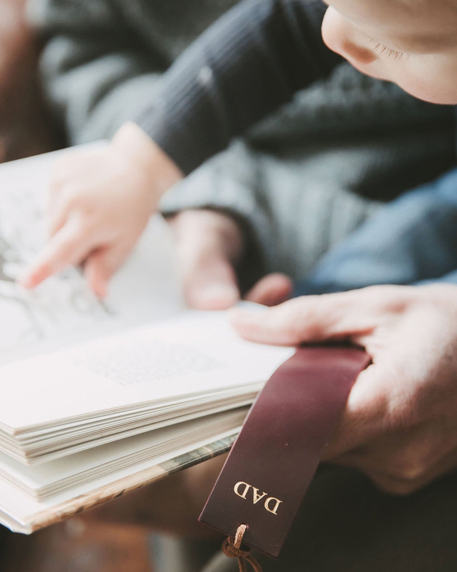 Brown leather bookmark with "DAD" on, held by a father and baby whilst reading a book.