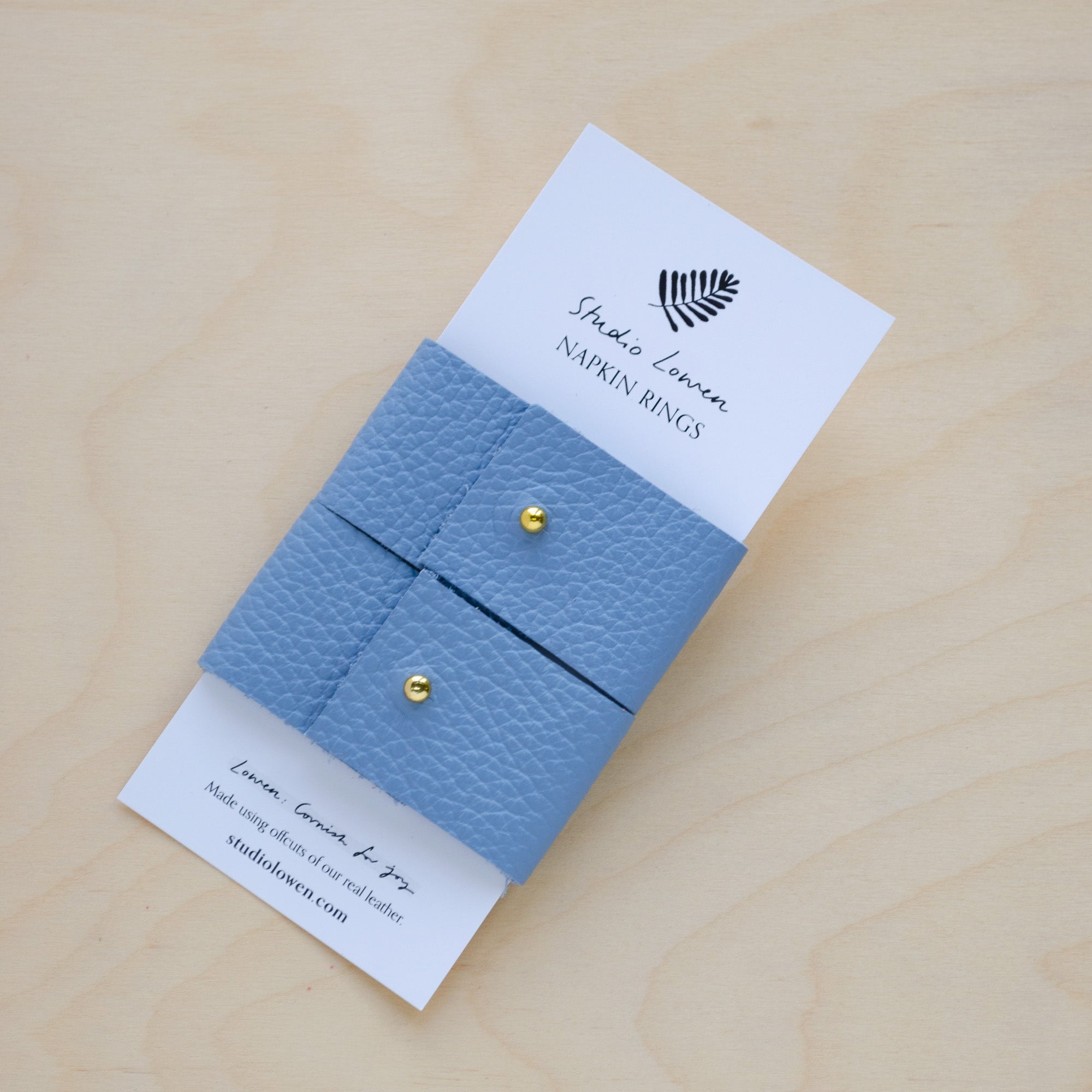 Pair of soft Periwinkle blue leather napkin rings with a gold stud on a white card packaging.