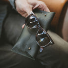 Dark Green leather glasses case in a man's hands with some brown Rayban sunglasses.